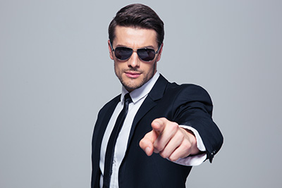 Handsome businessman in sunglasses pointing at camera over gray background